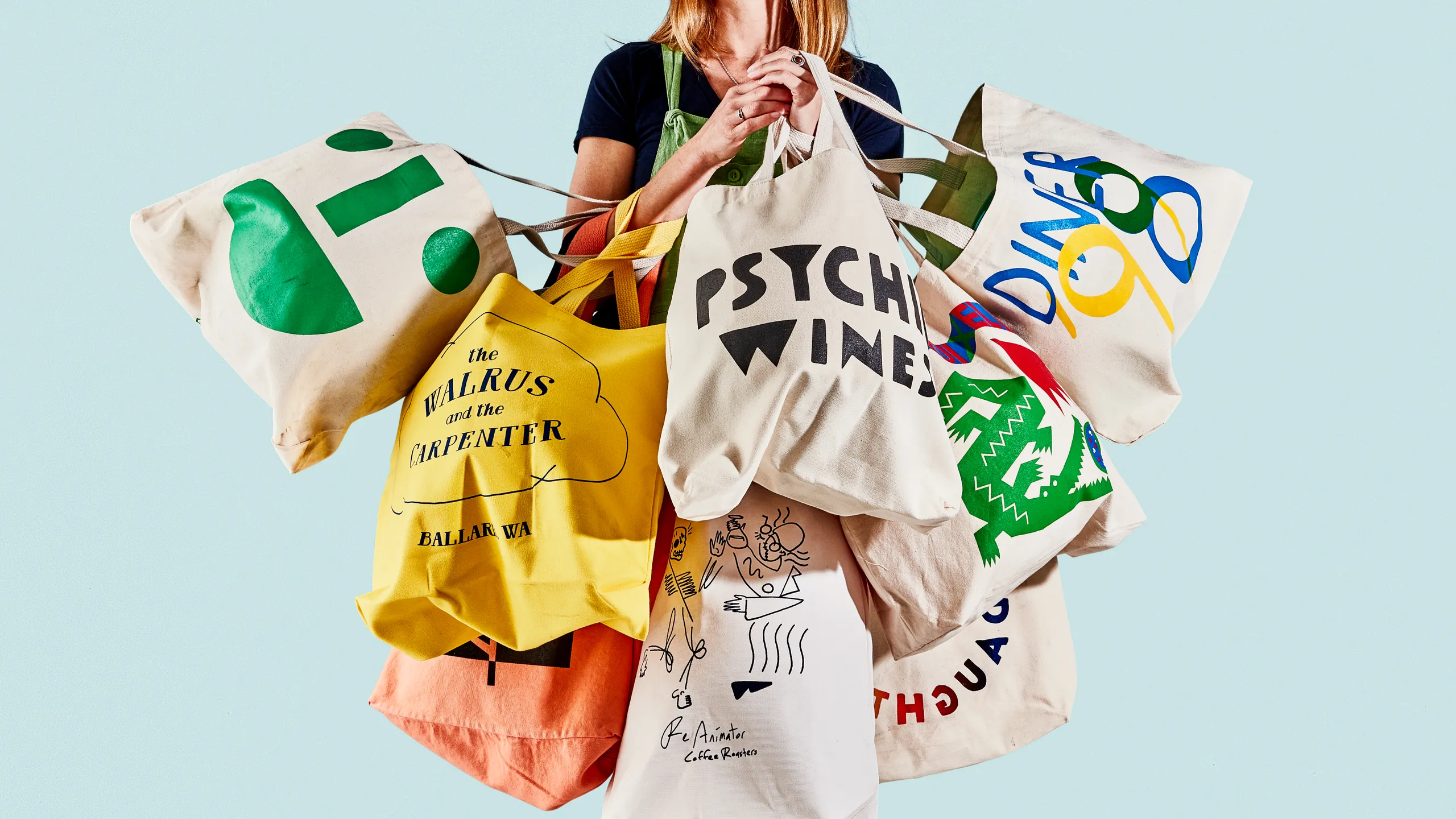 Person holding many printed canvas tote bags with colorful logos and designs