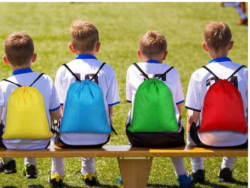 Four young soccer players sitting on a bench with colorful drawstring bags in yellow, blue, green, and red