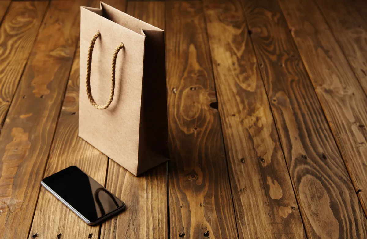 Eco-friendly paper bag with rope handle beside smartphone on wooden table