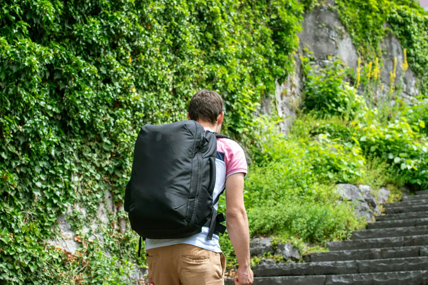 Man with black backpack walking up stone steps beside green ivy wall
