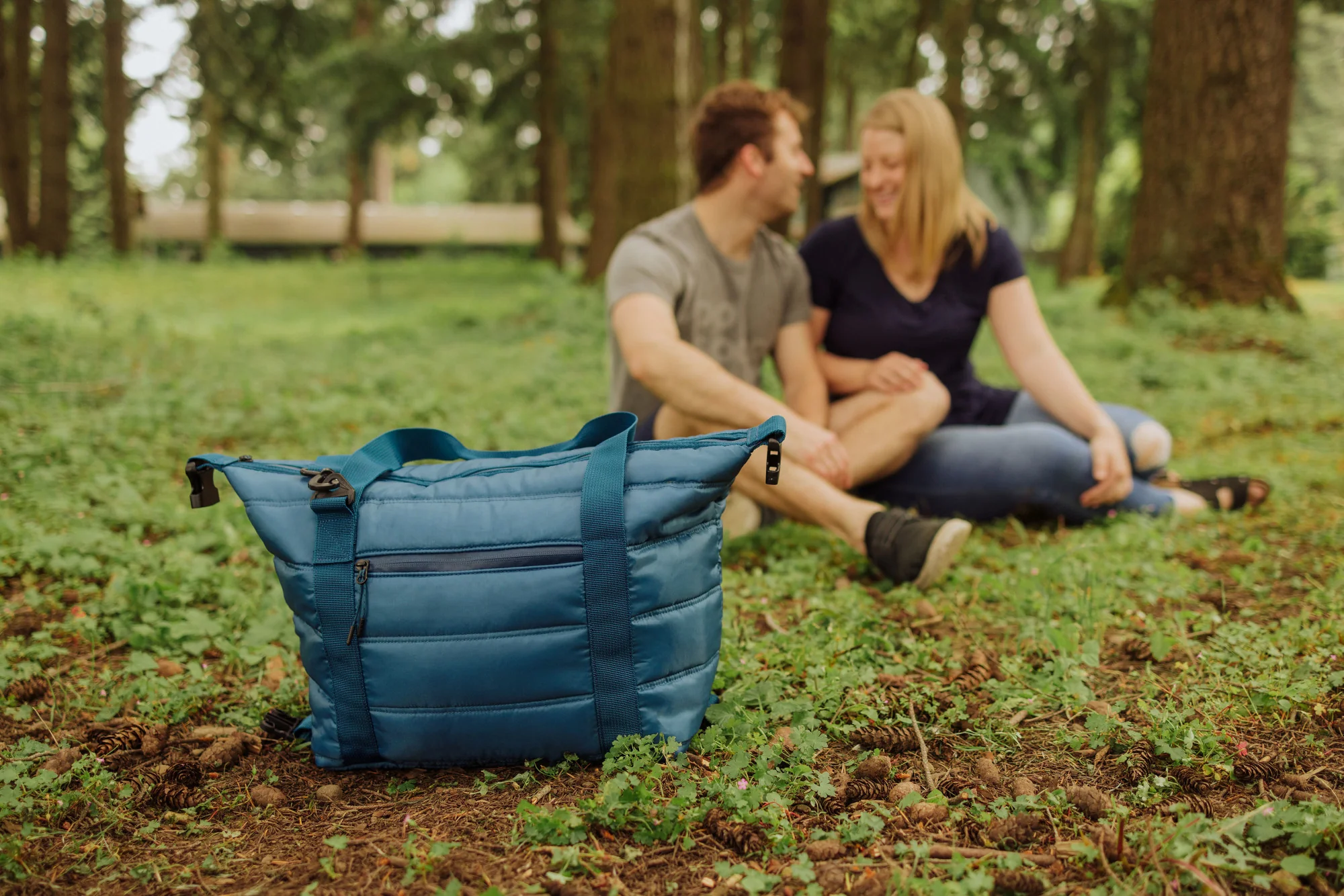 Blue insulated tote bag placed on forest ground near smiling couple