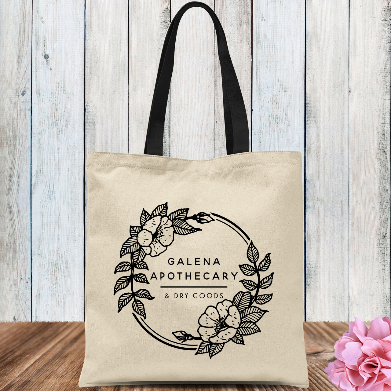 Beige tote bag with floral Galena Apothecary logo and black handles