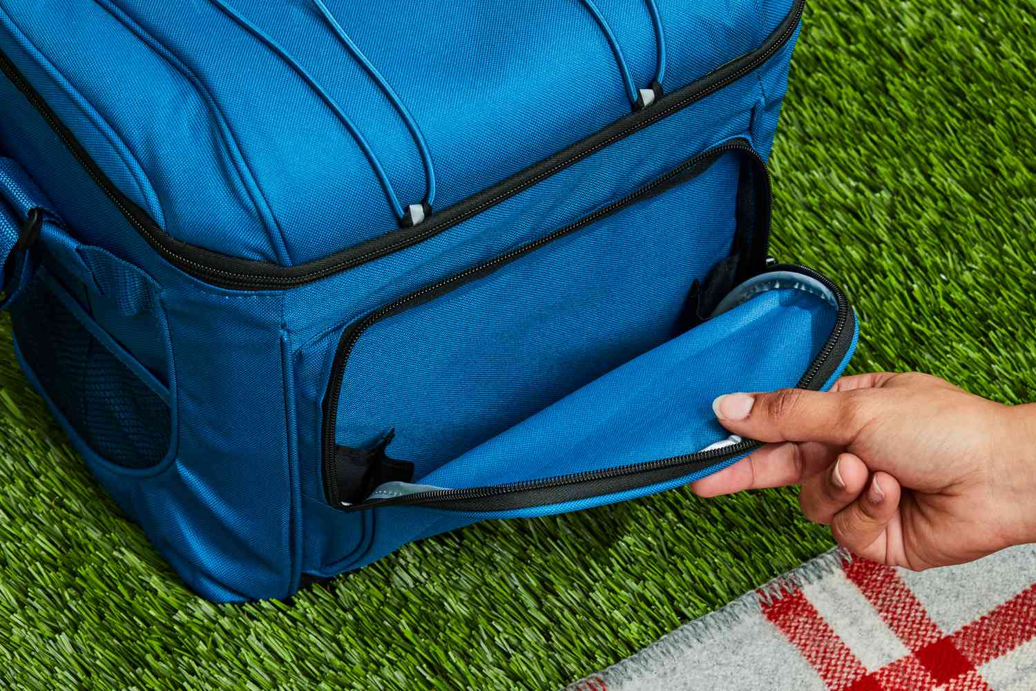 Hand opening front pocket of a blue insulated cooler bag on grass